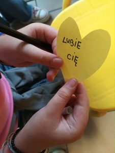Wycięte z żółtej kartki serce z napisem lubie Cię. Serce trzymane w dłoniach uczennicy.